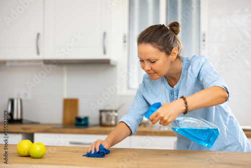Attentive woman uses spray while wiping the tabletop with a rag in a modern kitchen at home