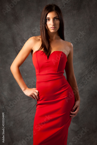 Portrait of young woman in red dress, standing and posing with hand on hip, looking at camera, grey background photo