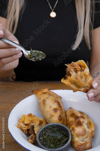 Eating empanadas at the restaurant. Closeup view of a woman hands pouring spicy chimichurri sauce with a spoon into a meat empanada.