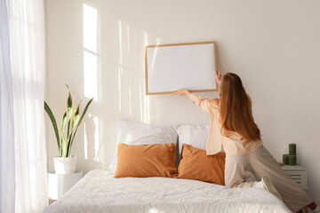 Young woman hanging blank frame on light wall in bedroom, back view