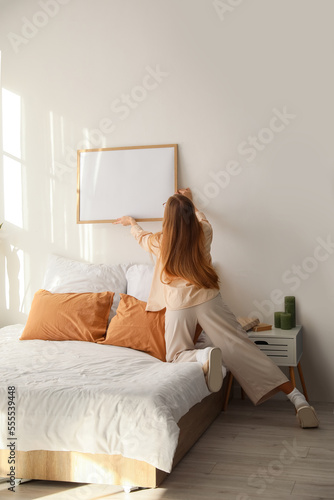 Young woman hanging blank frame on light wall in bedroom, back view