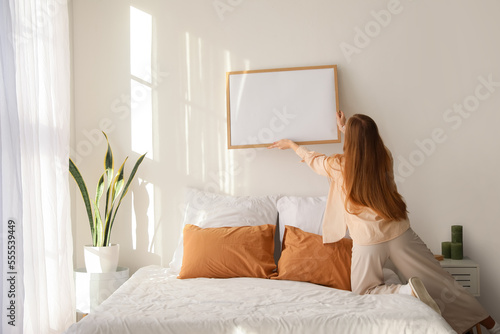 Young woman hanging blank frame on light wall in bedroom, back view photo