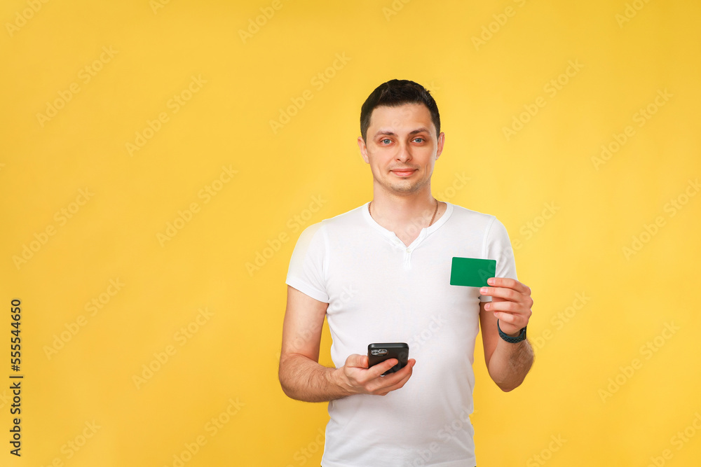 handsome young man holding a cellphone and a bank card in his hand to pay for purchases on the Internet in a white t-shirt on a yellow background. handsome young man holding a cellphone and a bank card in his hand to pay for purchases on the Internet in a white t-shirt on a yellow background.