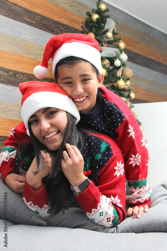 Single divorced mom and son Latino dressed in ugly sweters play in bed next to the Christmas tree with Santa hats give each other hugs and kisses show their love in solitude Single divorced mom and son Latino dressed in ugly sweters play in bed next to the Christmas tree with Santa hats give each other hugs and kisses show their love in solitude