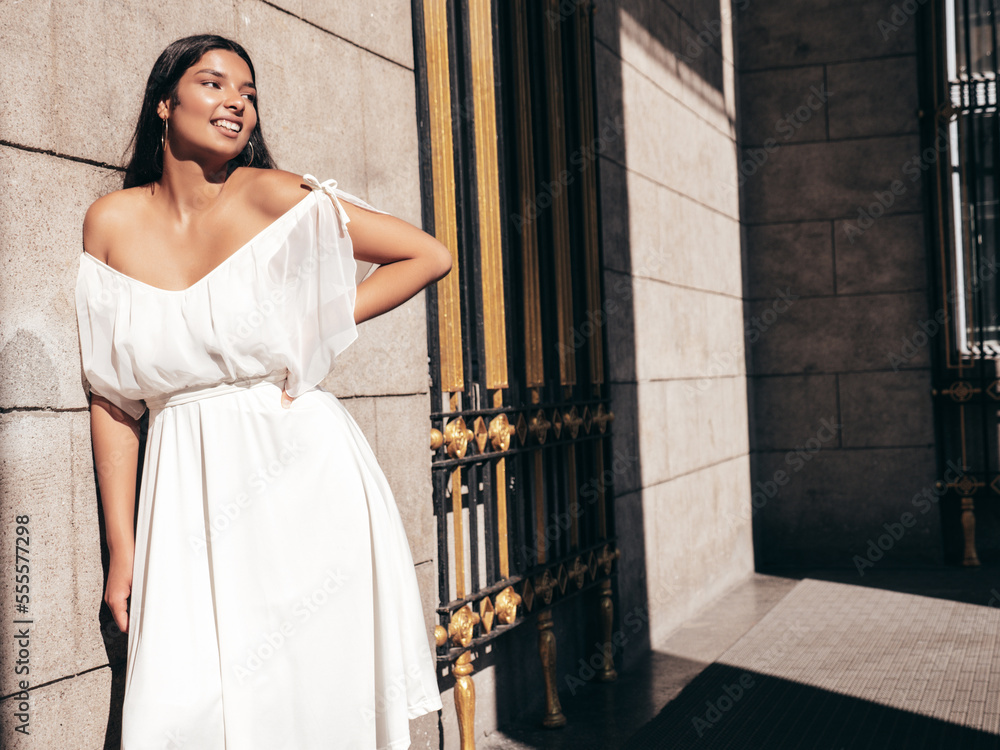 Fototapeta premium Young beautiful smiling hipster woman in white dress. Sexy carefree model posing on the street background at sunset. Positive female outdoors at warm sunny day. Cheerful and happy