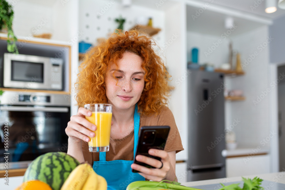Smiling pretty woman looking at mobile phone and holding glass of orange juice while cooking fresh vegetables in kitchen interior at home Smiling pretty woman looking at mobile phone and holding glass of orange juice while cooking fresh vegetables in kitchen interior at home