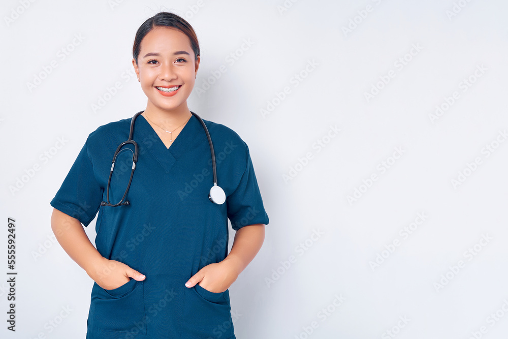 Smiling young Asian woman nurse wearing blue uniform with stethoscope holding hands in pockets isolated on white background. Healthcare medicine concept Smiling young Asian woman nurse wearing blue uniform with stethoscope holding hands in pockets isolated on white background. Healthcare medicine concept