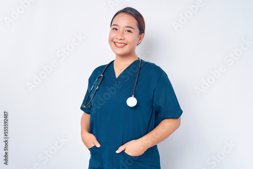 Smiling young Asian woman nurse wearing blue uniform with stethoscope holding hands in pockets isolated on white background. Healthcare medicine concept