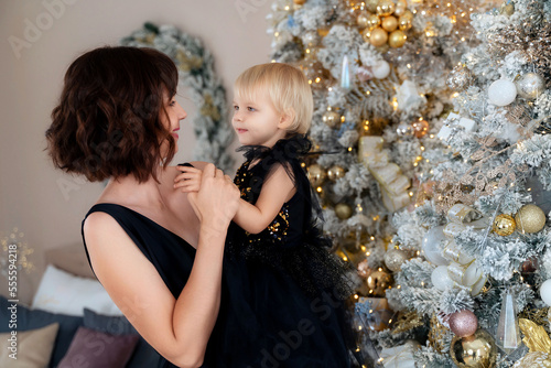 Mother with her daughter 2 years old near the Christmas tree. Both are dressed in black dresses, the mother holds the girl in her arms and both look at each other. The family celebrates Christmas.