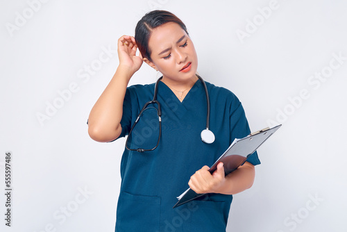Confused young Asian woman nurse wearing a blue uniform with a stethoscope looking at a clipboard isolated on white background. Healthcare medicine concept
