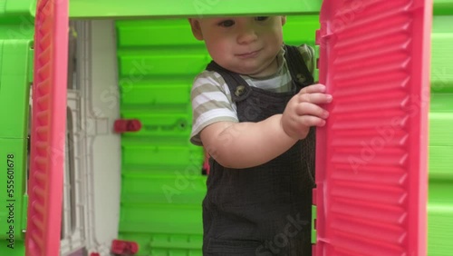 Childhood, toddler, motherhood, summer. Happy smiling Baby Boy Kid playing hide-and-seek and looks out the window in plastic playhouse. Little active child plays outside in the garden Playground