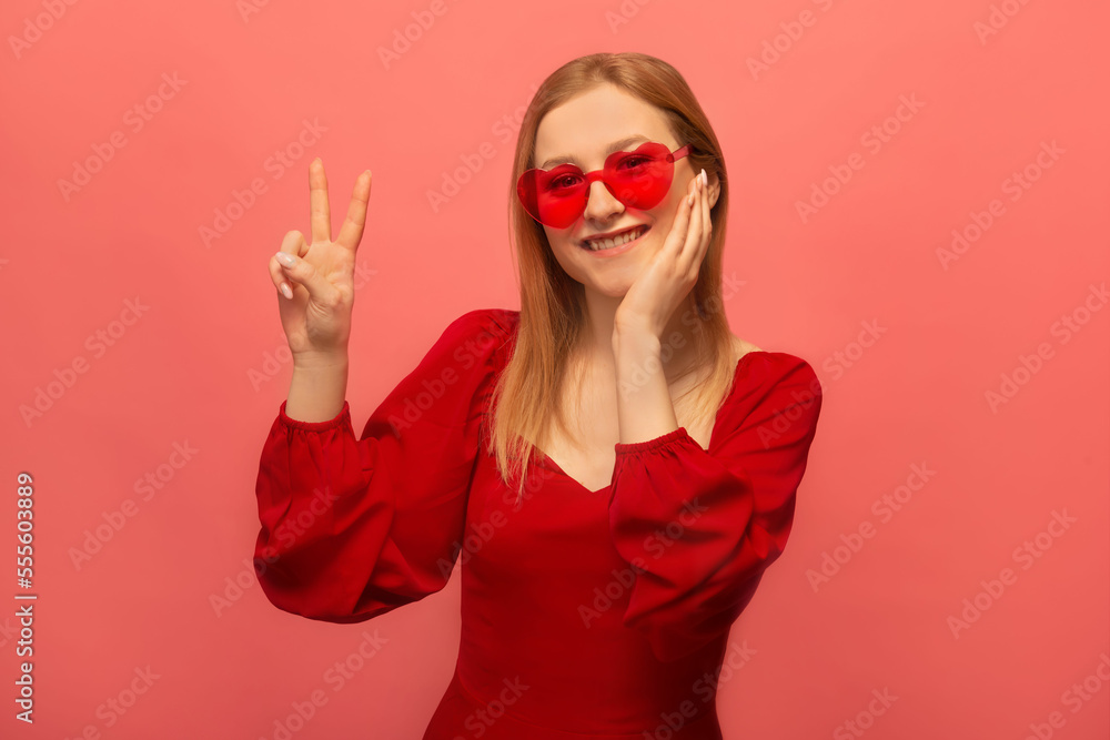 Stylish happy smiling young woman in red dress, french beret and trendy heart shaped glasses showing peace gesture v sign with two fingers and keeping hand on face isolated on colored pink background. Stylish happy smiling young woman in red dress, french beret and trendy heart shaped glasses showing peace gesture v sign with two fingers and keeping hand on face isolated on colored pink background.