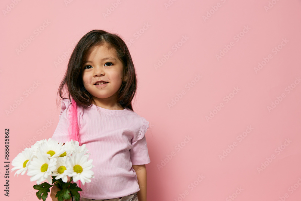 a little girl of preschool age stands on a pink background in a pink T-shirt and poses with a bouquet of white daisies and stretches them forward smiling shyly looking away a little girl of preschool age stands on a pink background in a pink T-shirt and poses with a bouquet of white daisies and stretches them forward smiling shyly looking away