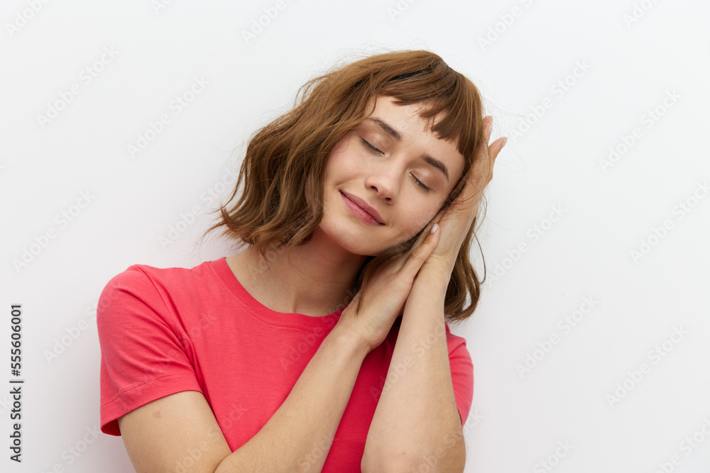 Obraz premium a gentle, relaxed, sleepy woman stands in a red T-shirt on a white background and holds her hands folded in her palms under her head, closing her eyes