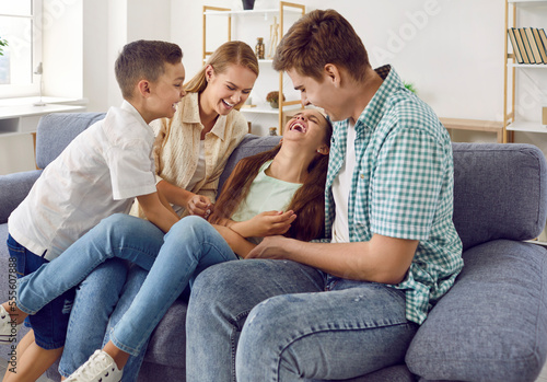 Happy family are playing together, they are tickling a girl sitting on couch and laugh in living room. Mom, dad, son and daughter spending time together. Family lifestyle, love, fun and care concept.