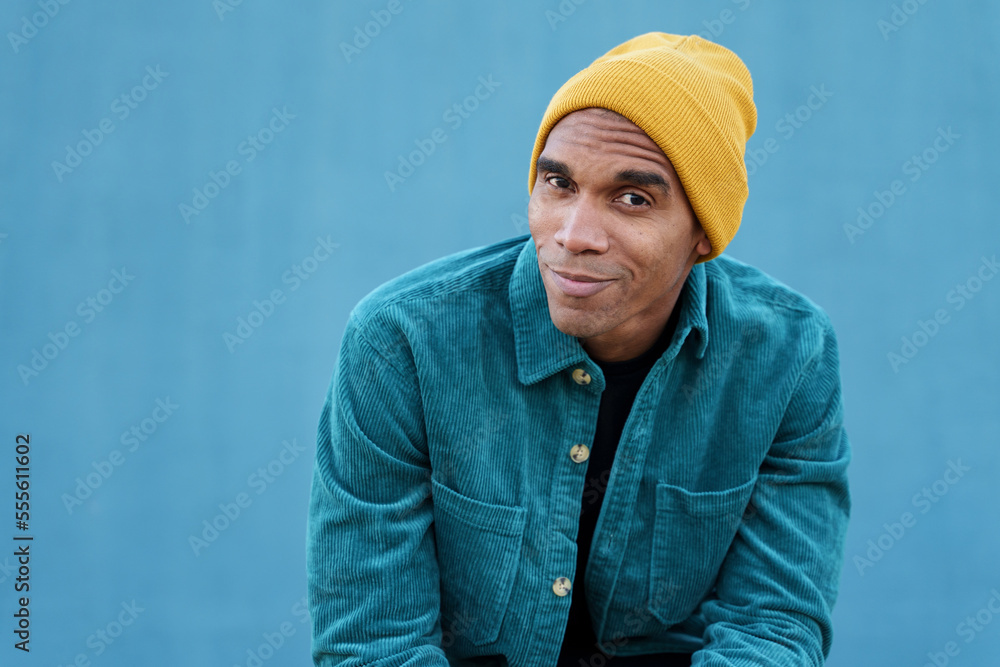Positive African American man in yellow beanie and blue jacket sitting against blue wall Positive African American man in yellow beanie and blue jacket sitting against blue wall