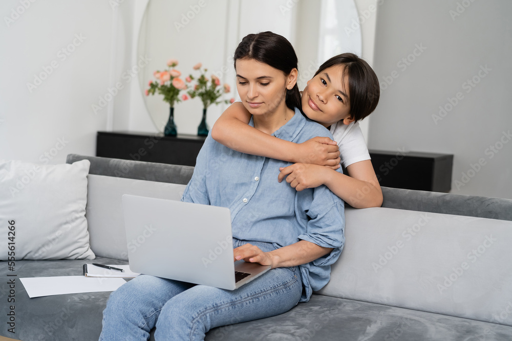 Smiling asian child hugging mom using laptop in living room Smiling asian child hugging mom using laptop in living room