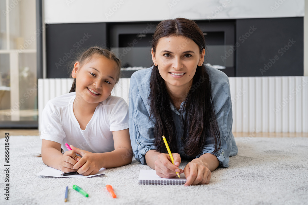 Smiling woman writing on notebook and looking at camera near asian daughter with markers at home Smiling woman writing on notebook and looking at camera near asian daughter with markers at home