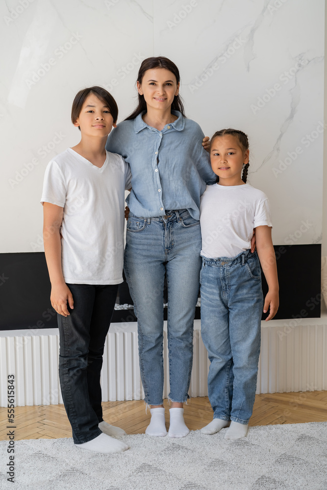 Smiling woman hugging asian kids and looking at camera at home Smiling woman hugging asian kids and looking at camera at home