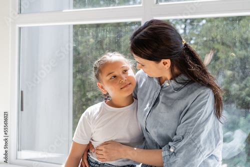 Woman hugging asian daughter near window at home