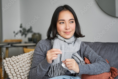 Smiling asian woman holding thermometer and looking pleased, feeling better after cold, got rid of fever, has normal temperature