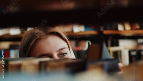Young caucasian woman student standing between bookcases and thoroughly choosing a book used in her education from the shelf in the library close up