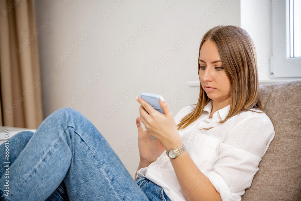 Home affairs room woman telephone. White cozy bed and a beautiful girl in a white shirt and jeans, reads messages on the phone, the concept of home and comfort. Against the background of the living Home affairs room woman telephone. White cozy bed and a beautiful girl in a white shirt and jeans, reads messages on the phone, the concept of home and comfort. Against the background of the living
