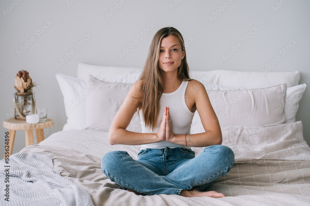 Focused young adult caucasian woman in white t-shirt sitting on bed in yoga meditation pose with folded hands looks at camera. Confident Hispanic female makes breath exercise at home. Focused young adult caucasian woman in white t-shirt sitting on bed in yoga meditation pose with folded hands looks at camera. Confident Hispanic female makes breath exercise at home.