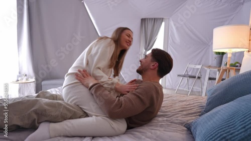 Happy cheerful young couple cuddling on bed laughing out loud. Side view portrait of loving Caucasian boyfriend and girlfriend having fun in bedroom indoors. Happiness and love concept