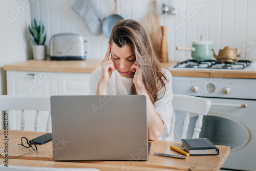 Exhausted young brunette caucasian woman in white t-shirt sitting at kitchen desk with laptop touches temples eyes closed feels fatigue, headache. Overloaded hispanic girl works at home. Tired student
