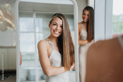 Gorgeous caucasian woman with long hair standing at mirror smiles wide looks at camera satisfied by healthy skin and hair. Beautiful Italian female in underwear happy after hair care procedure.