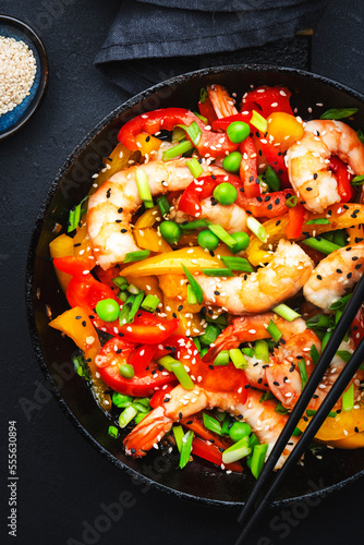 Stir fry with shrimps, red and yellow paprika, green pea, chives and sesame seeds in frying pan. Asian cuisine dish. Black stone kitchen table background, top view