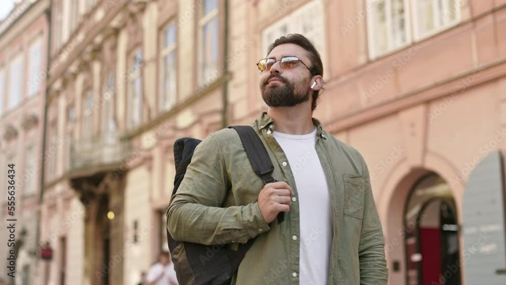 Side view of male tourist standing on street, admiring buildings, holding, using smartphone. Handsome man with beard and earphones traveling, wearing sunglasses. Concept of tourism. Side view of male tourist standing on street, admiring buildings, holding, using smartphone. Handsome man with beard and earphones traveling, wearing sunglasses. Concept of tourism.