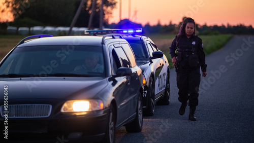Highway Traffic Patrol Car Pulls over Vehicle on Road. Male Police Officer Approaches Driver Asks for License and Registration, Black Female Colleague Covers Him. Cinematic Dusk Wide Shot