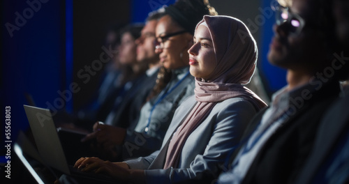 Arab Female Sitting in a Dark Crowded Auditorium at a Human Rights Conference. Young Muslim Woman Using Laptop Computer. Activist in Hijab Listening to Inspiring Speech About Global Initiative. photo