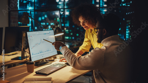 Team of Two Young Entrepreneurs Talking, Discussing Growth Strategy at a Desk with Computer. Stylish Businesspeople Work on an Investment and Marketing Report in a Creative Agency in the Evening.