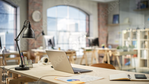 Charging Laptop on a Working Station in a Bright Spacious Empty Workplace. Office Supplies Are on the Table. Big Windows with City Buildings View in the Background.