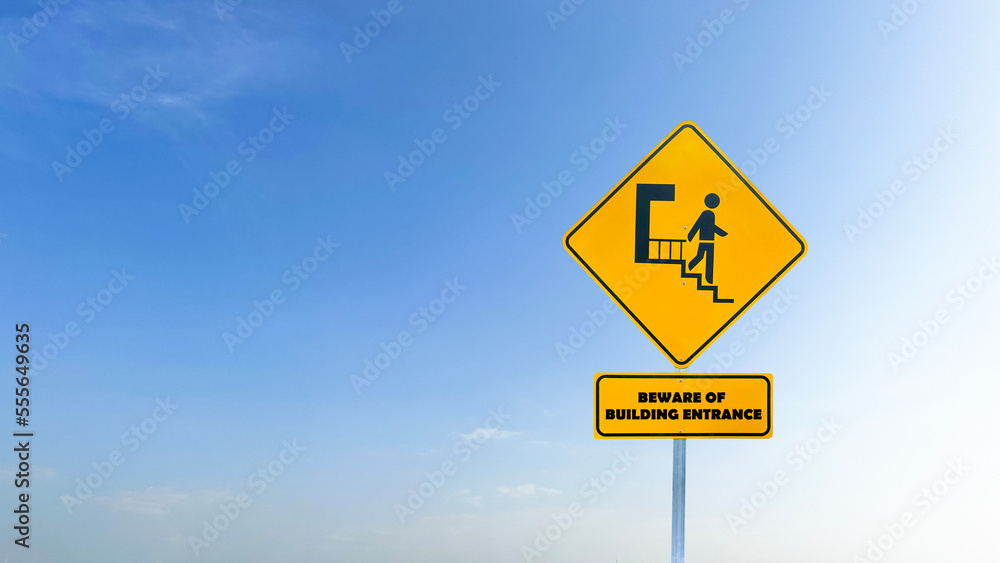 Yellow sign with graphic of person stepping down stairs and the text beware of building entrance on blue sky background