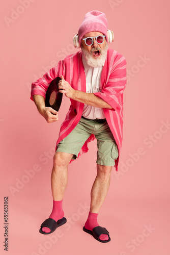 Portrait of stylish senior, old man in stylish bright outfit with knitted hat, sunglasses and headphones posing with vinyl over pink background