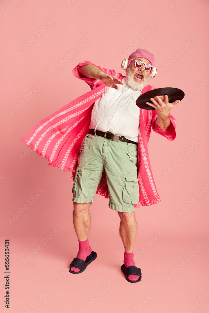 Portrait of stylish senior, old man in stylish bright outfit with vinyl posing over pink background. Club, dj