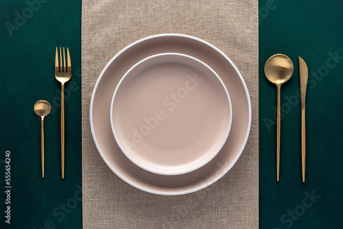 Festive place setting with beige napkin. Empty plates and gold cutlery on dark green background. Top view. Dining table in luxury restaurant. Card or menu template, flat design. Tableware, crockery.