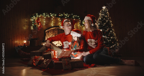 Caucasian family of three sitting in decorated room near christmas tree, little girl opening her christmas gift with something special - christmas spirit, togetherness concept 