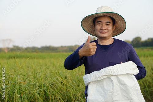 Handsome Asian man farmer is at paddy field, wears hat, holds white sack of organic fertilizer, thumbs up, confident. Concept, farmer satisfied in product for agriculture. 
