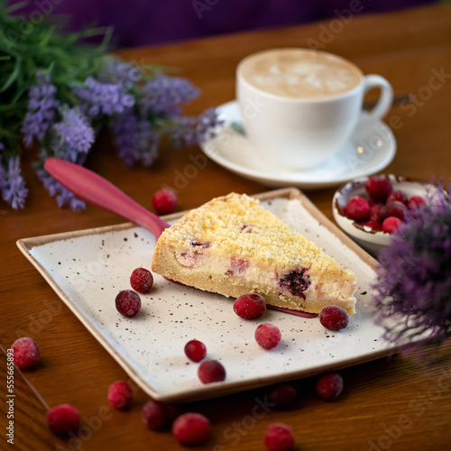 Portion of cheesecake on dessert spatula on plate. Cheesecake with berries and cup of coffee on table. Bouquet of lavender and scattering of berries on wooden table. Still life. 