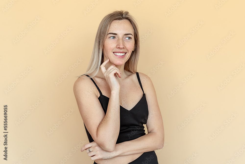 Dreamy pretty young woman with freckles touch chin thought choose decide solve problems dilemmas, wear fashionable top, stands over beige background. Thinking positive concept