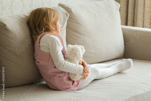 Foto Sad little girl holding white teddy bear and sitting on sofa at living room