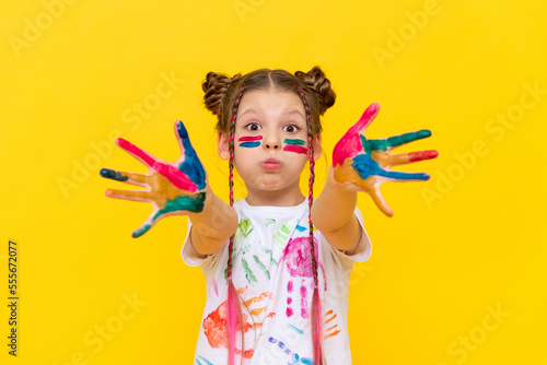 Portrait of a little girl smeared in multicolored paints for drawing on a yellow isolated background. The schoolgirl puffed out her cheeks. Development of children's creativity and art.