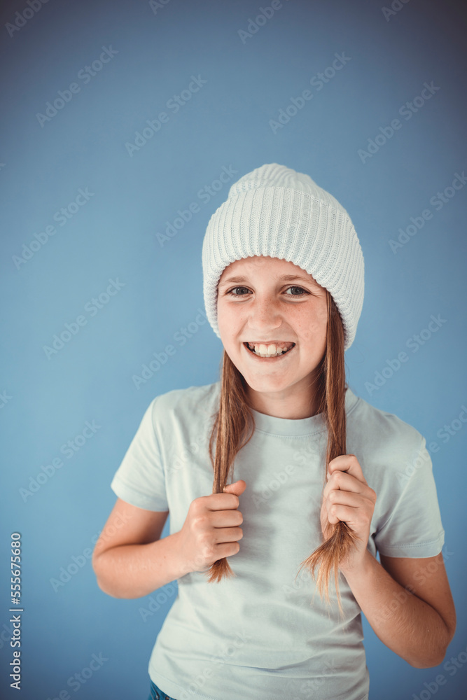 cool and pretty girl with winter wool beanie is standing in front of blue background and making grimaces, faces