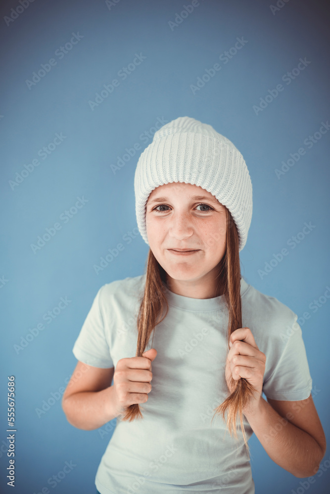 cool and pretty girl with winter wool beanie is standing in front of blue background and making grimaces, faces