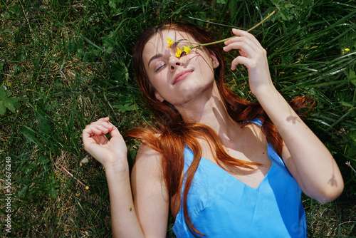Woman smile with teeth lying on the green grass in a blue dress in the spring sunshine with yellow flowers, happiness, red long hair